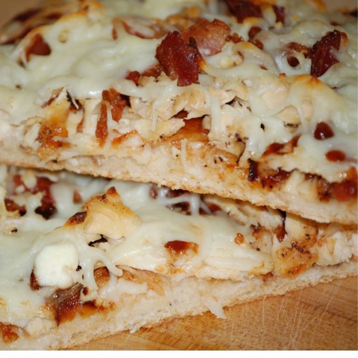 Two thick slices of pizza topped with shredded chicken, crispy bacon, melted mozzarella cheese, and barbecue sauce on a wooden surface.