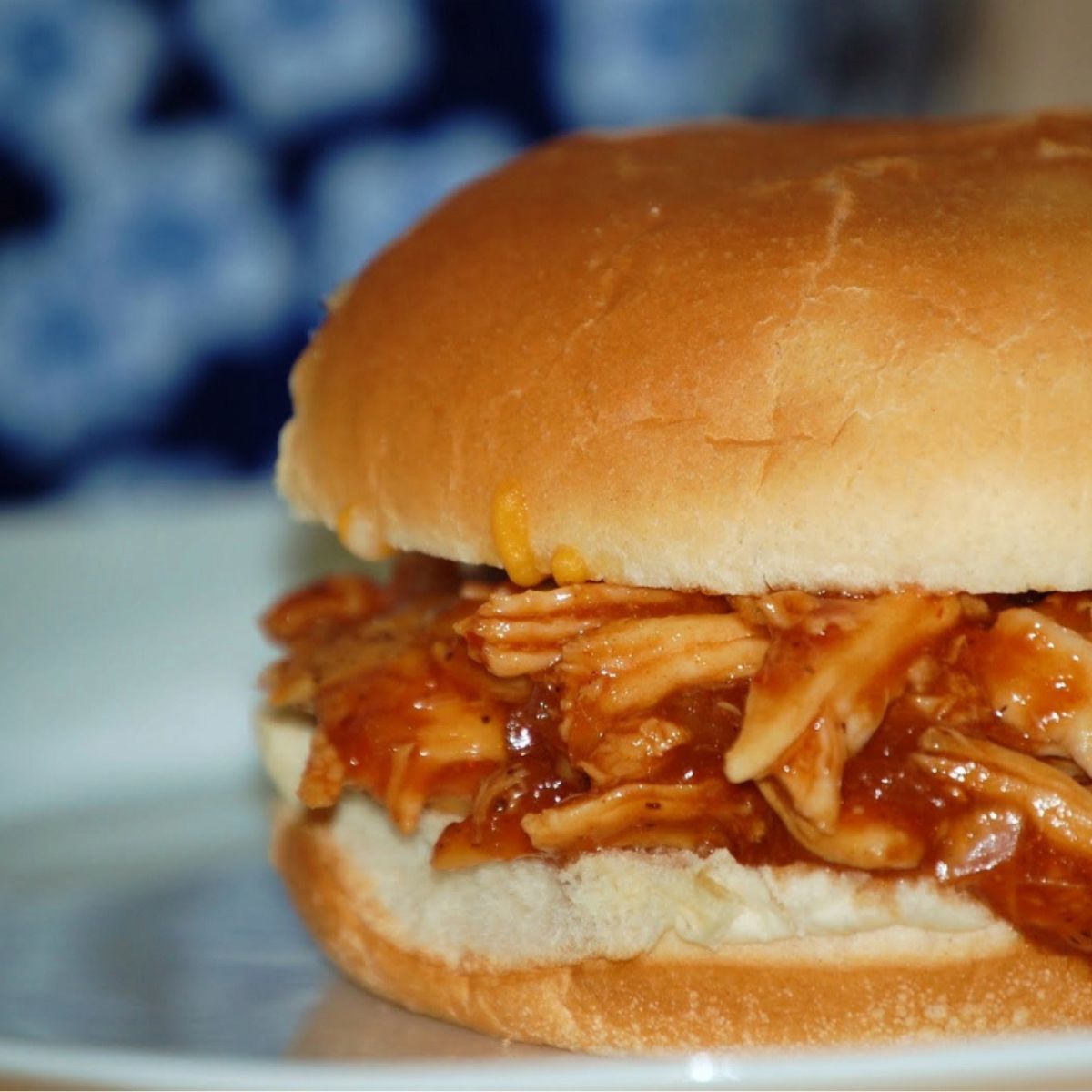 Pulled barbecue chicken piled inside a soft sandwich bun, with tangy sauce visible on the chicken and a blue patterned background.