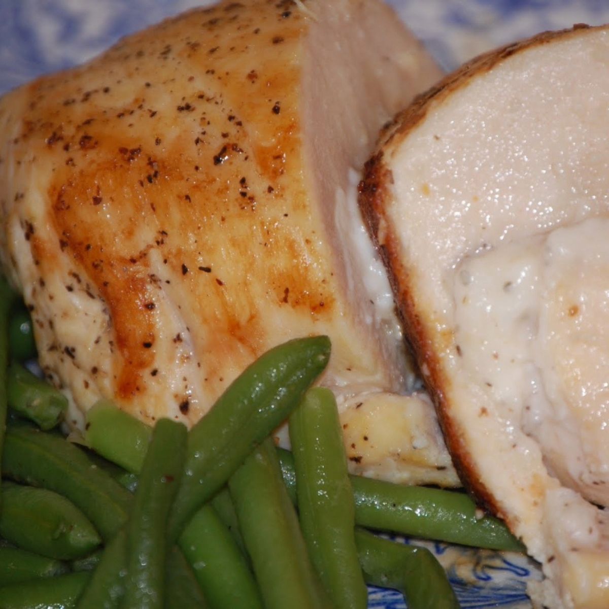Roasted chicken breast sliced to show a cheesy filling, served alongside crisp cooked green beans on a patterned blue plate