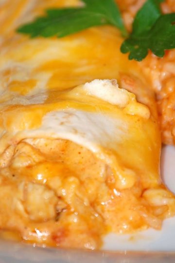 Cheesy chicken enchilada covered in melted orange and white cheese, served with a portion of Mexican-style rice and a garnish of parsley.