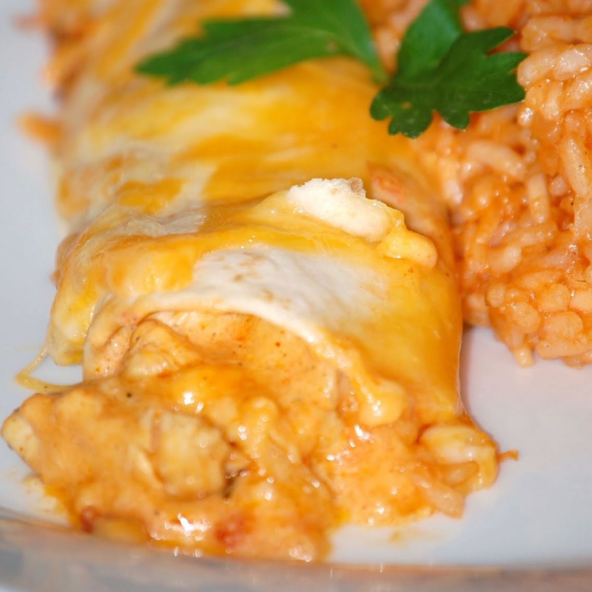 Cheesy chicken enchilada covered in melted orange and white cheese, served with a portion of Mexican-style rice and a garnish of parsley.