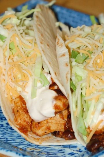 Two soft flour tortillas filled with seasoned chicken pieces, shredded lettuce, shredded cheddar cheese, and a creamy white sauce, served on a blue patterned plate with a small dish of sauce on the side.
