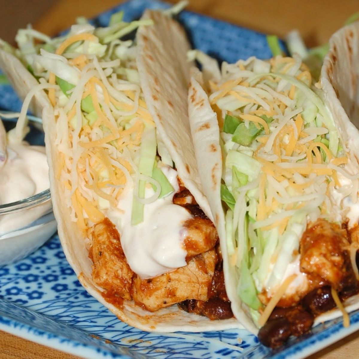 Two soft flour tortillas filled with seasoned chicken pieces, shredded lettuce, shredded cheddar cheese, and a creamy white sauce, served on a blue patterned plate with a small dish of sauce on the side.