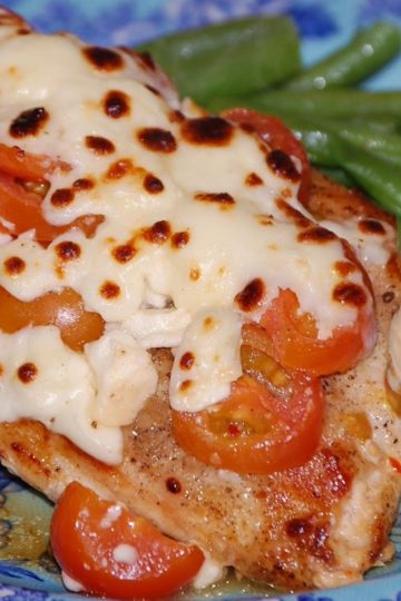 Chicken breast topped with melted mozzarella and feta cheese, sliced cherry tomatoes, and served with a side of cooked green beans on a blue floral plate.