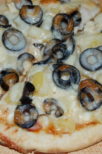 Pizza topped with melted mozzarella cheese, sliced black olives, and chunks of pineapple, resting on a wooden surface.