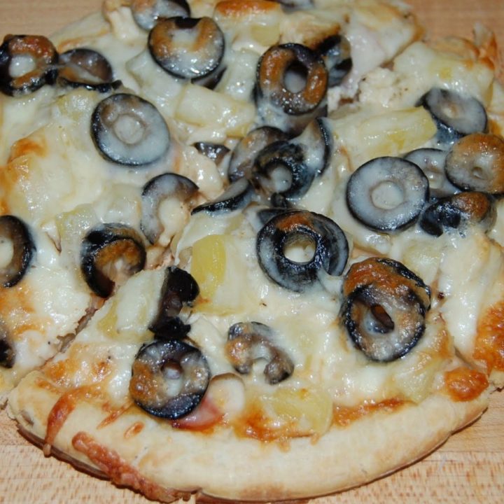 Pizza topped with melted mozzarella cheese, sliced black olives, and chunks of pineapple, resting on a wooden surface.