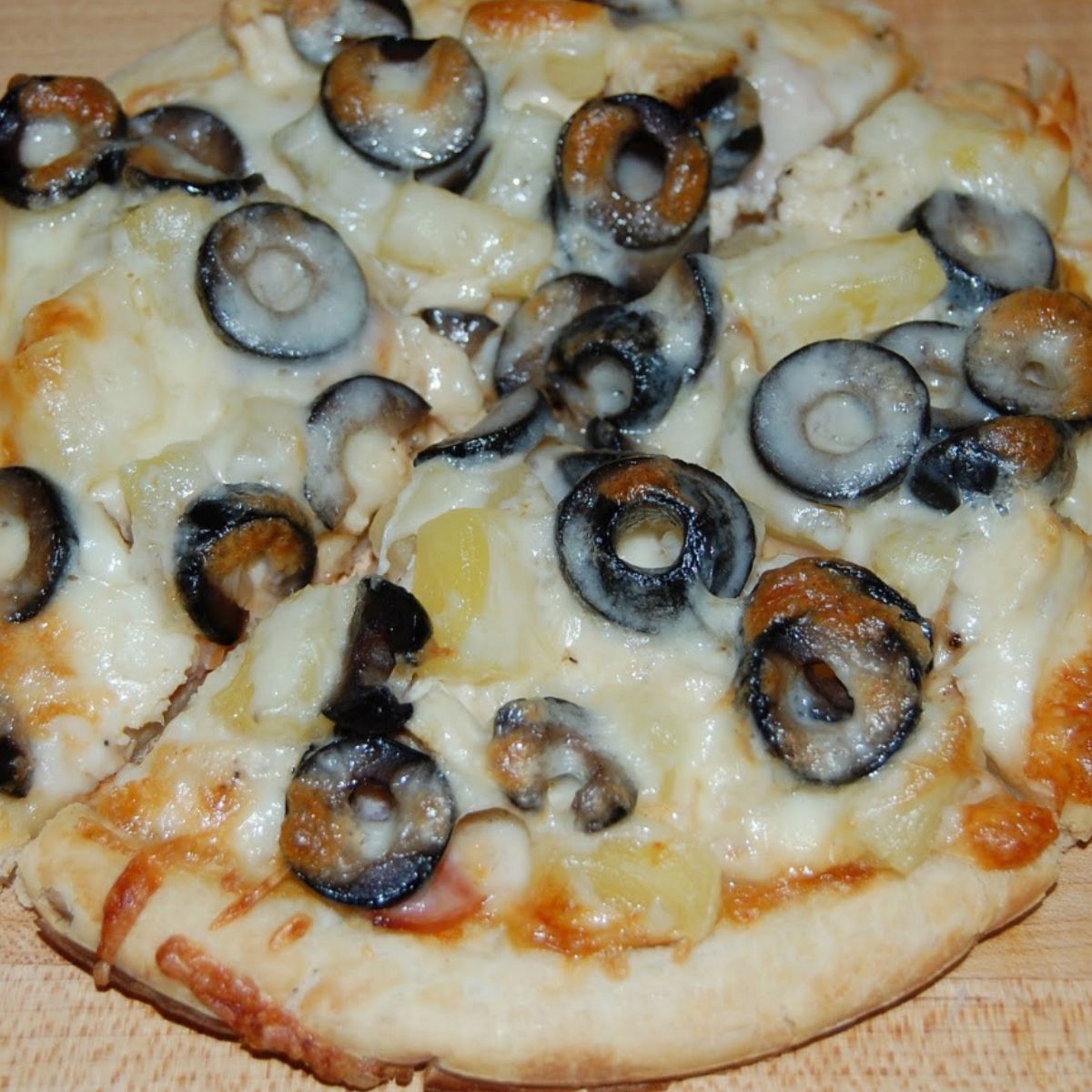 Pizza topped with melted mozzarella cheese, sliced black olives, and chunks of pineapple, resting on a wooden surface.