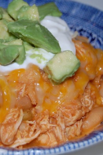 Bowl of shredded chicken and white beans in a chili sauce, topped with melted cheese, diced avocado, and a dollop of sour cream.