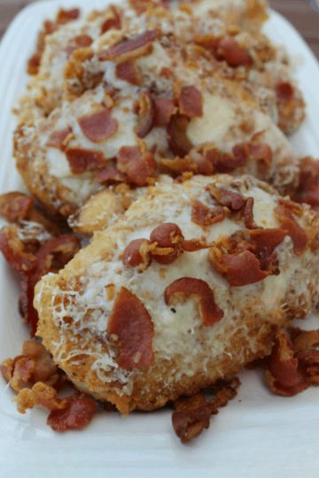 Golden-brown breaded chicken breasts topped with melted asiago cheese and crispy bacon pieces, served on a white platter.